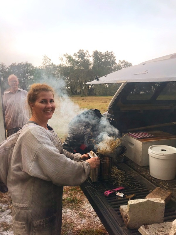 Photo from the Tampa Bees course page of an instructor in a bee suit lighting a smoker beside a truck bed, with a man behind her. An instructor photo, not a logo.