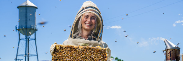 A smiling beekeeper in a white veil holds a frame of comb under a blue sky, from the University of Montana Beekeeping program banner.